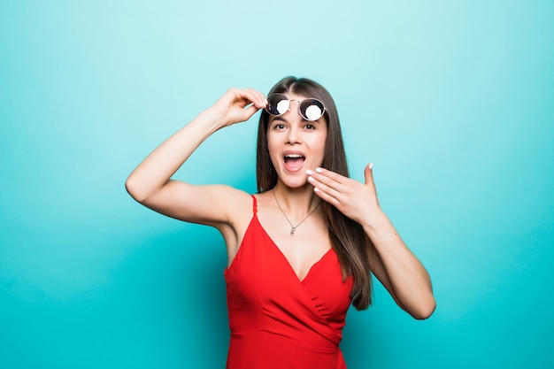 Geschokt mooie jonge vrouw in rode minikleding en zonnebril bedekken mond met hand op blauwe muur.