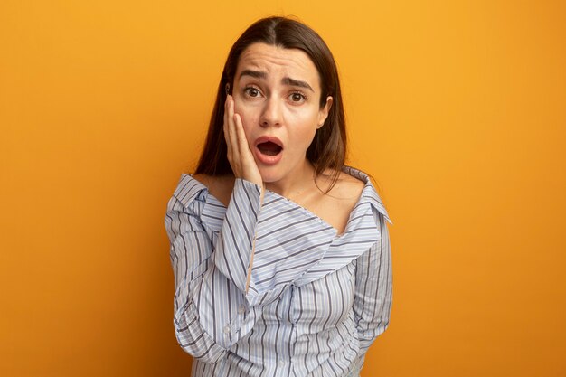 Geschokt mooie vrouw legt hand op gezicht geïsoleerd op oranje muur