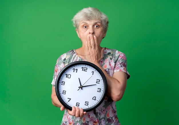 Geschokt oudere vrouw legt hand op de klok van de mondholding die op groene muur wordt geïsoleerd