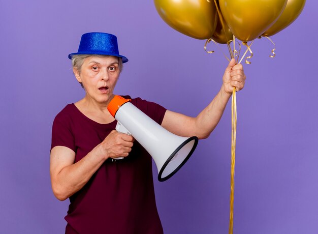 Geschokt oudere vrouw met feestmuts houdt helium ballonnen en luidspreker geïsoleerd op paarse muur met kopie ruimte