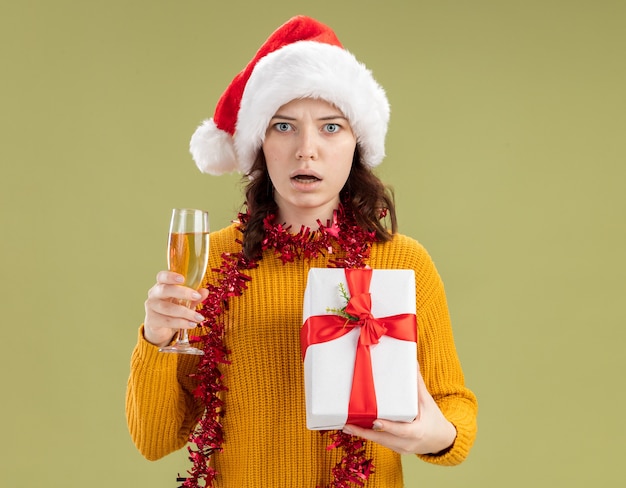 Geschokt slavisch meisje met kerstmuts en met slinger om nek met glas champagne en kerstcadeaudoos geïsoleerd op olijfgroene muur met kopieerruimte