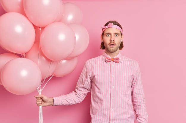 Geschokt verrast man houdt bos ballonnen hoort geweldig nieuws draagt hoofdband elegant shirt met strik viert iets gaande verjaardagsfeestje geïsoleerd over roze muur