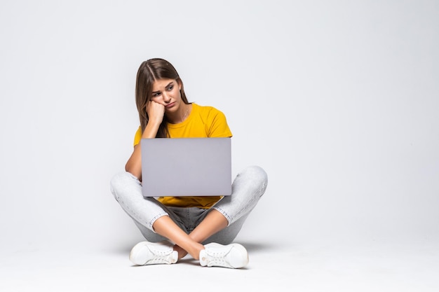 Geschokt verwarde vrouw in t-shirt zittend op de vloer met laptopcomputer op grijs