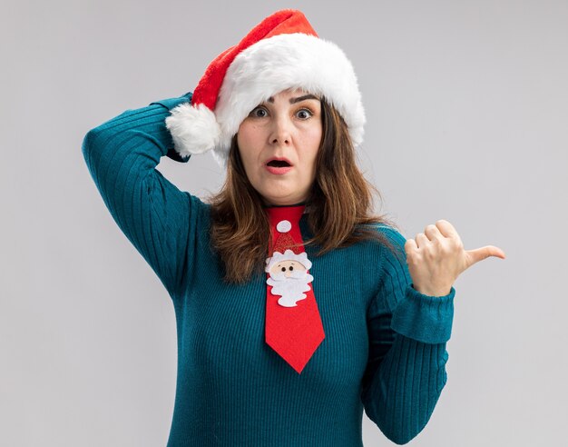 Geschokt volwassen blanke vrouw met kerstmuts en santa stropdas zet hand op het hoofd en wijst naar kant geïsoleerd op een witte achtergrond met kopie ruimte