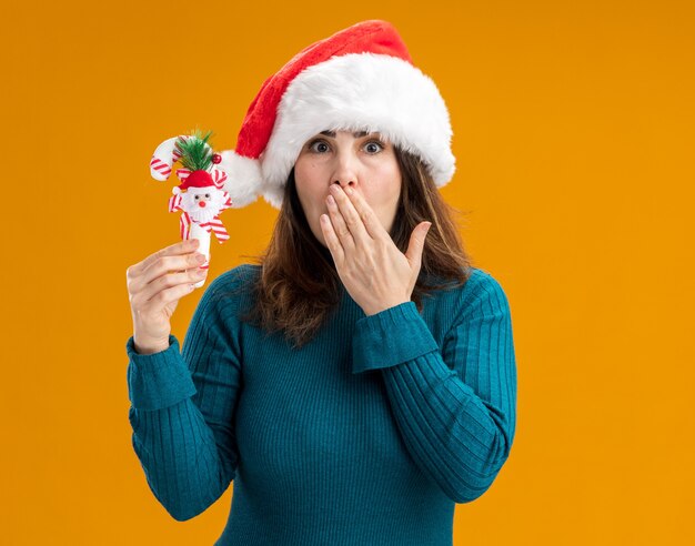 geschokt volwassen blanke vrouw met kerstmuts legt de hand op de mond en houdt snoepgoed geïsoleerd op een oranje achtergrond met kopie ruimte