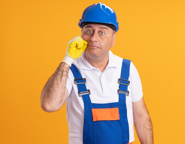 Gratis foto geschokt volwassen bouwer man in uniform dragen van beschermende handschoenen ritsen mond geïsoleerd op oranje muur