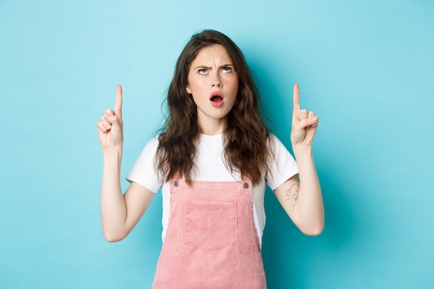 Geschokte beledigde jonge vrouw fronsend, hijgend en kijkend met een teleurgesteld en beledigd gezicht, iets respectloos tonend, staande tegen een blauwe achtergrond.