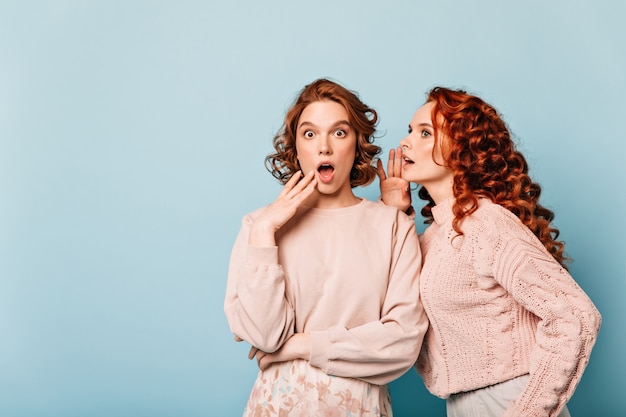 Geschokte dames die geruchten delen. Studio shot van verbaasde roddelmeisjes die zich voordeed op blauwe achtergrond.