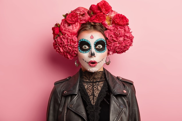 Geschokte, doodsbange jonge vrouw heeft een eng spookgezicht, draagt artistieke make-up voor Day of Dead-vakantie, draagt een zwart leren jack, modellen op een roze studioachtergrond. Schedel vrouwtje symboliseert de dood