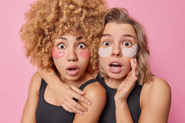 Geschokte emotionele vrouwen omarmen en staren afgeluisterde ogen naar de camera passen schoonheidshydrogelpleisters toe voor het verminderen van rimpels gekleed in casual t-shirts geïsoleerd op roze achtergrond Huidverzorgingsconcept