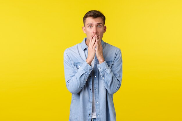 Geschokte en bezorgde blonde man die bezorgd naar de camera kijkt, lippen aanraakt en angstig naar de camera staart, hoort over een vreselijk ongeluk, medeleven en empathie uitdrukt, gele achtergrond.