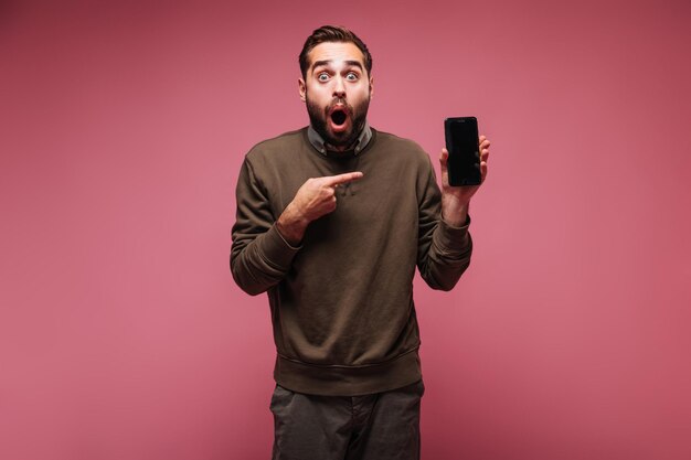 Geschokte man in donkere outfit pronkt met smartphone op roze achtergrond