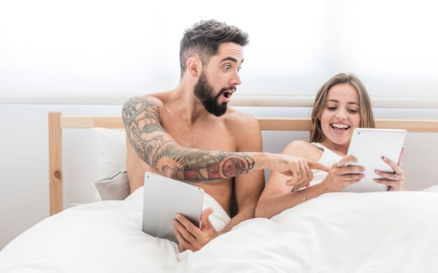 Geschokte mens die zijn gelukkige vrouw bekijkt die digitale tablet op bed gebruikt