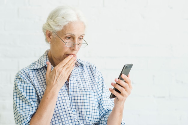 Geschokte oude vrouw die mobiel tegen achtergrond bekijkt