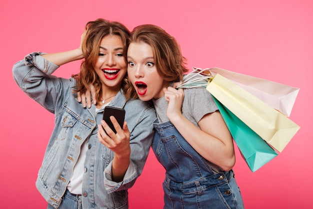 Geschokte twee vrouwenvrienden die het winkelen zakken houden gebruikend mobiele telefoon.