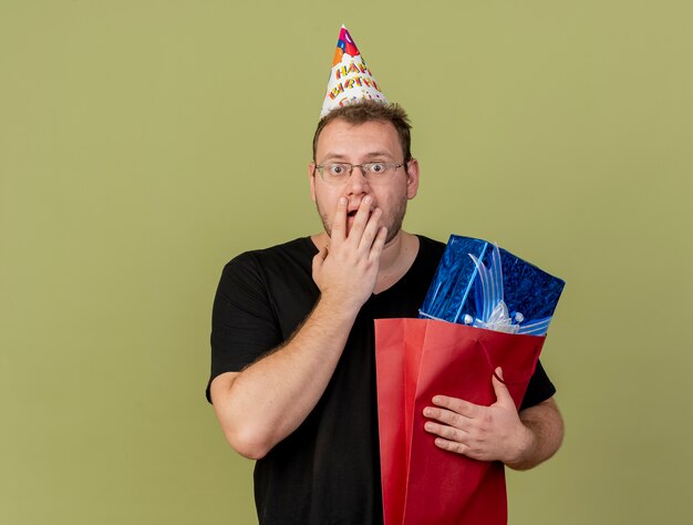 Geschokte volwassen Slavische man met optische bril met verjaardagspet legt hand op mond en houdt geschenkdoos in papieren boodschappentas