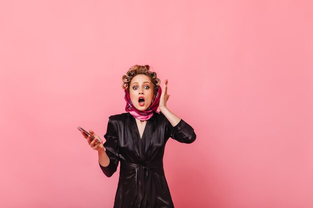 Geschokte vrouw die in sjaal smartphone op roze muur houdt