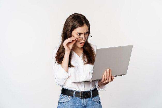 Geschokte vrouw die verward naar het scherm van de laptop kijkt, verbijsterd over iets op de computer, staande op een witte achtergrond
