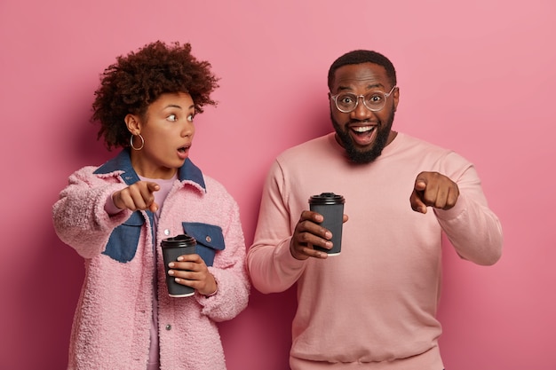 Geschokte vrouw en gelukkige man wijzen naar de camera, merken iets ongelooflijks op, drinken koffie uit wegwerpbekers, dragen stijlvolle outfit, drukken verschillende emoties uit