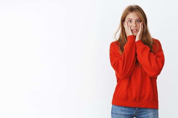 Geschokte vrouw kijkt met empathie naar vriend die vreselijk nieuws vertelt, handpalmen tegen de wangen drukt, tanden op elkaar klemt en naar adem snakkend bezorgd, nerveus en bezorgd, een schattige rode trui dragend