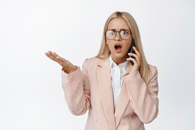 Geschokte zakenvrouw ontvangt slecht nieuws over de telefoon die haar schouders ophaalt en er gefrustreerd uitziet terwijl ze in een pak tegen een witte achtergrond staat