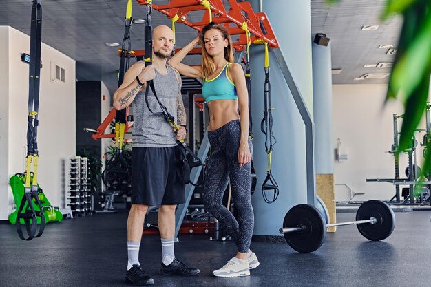 Geschoren hoofd atletische man en slanke brunette vrouw poseren in de buurt van trx bandjes staat in een sportschool club.