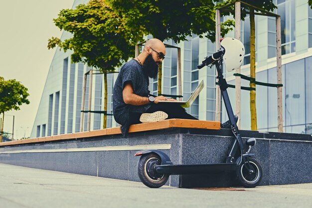 Geschoren hoofd getatoeëerd, bebaarde man met behulp van een laptop over modern gebouw achtergrond na de elektrische scooter rit.