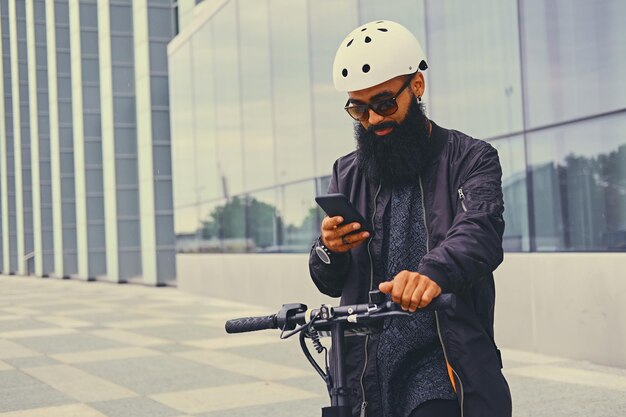 Geschoren hoofd getatoeëerd, bebaarde man met behulp van een smartphone over moderne gebouw achtergrond na de elektrische scooter rit.