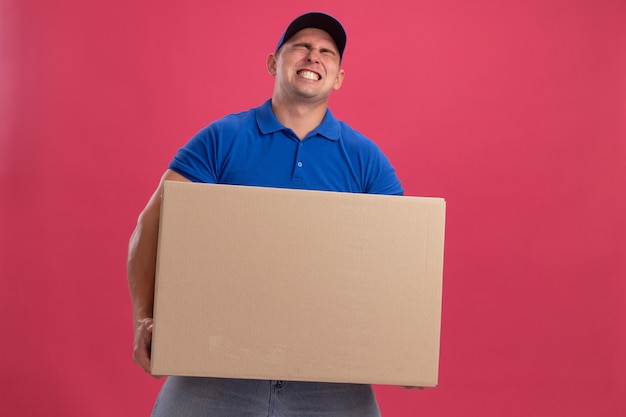 Gespannen jonge bezorger die uniform met pet draagt die grote doos houdt die op roze muur wordt geïsoleerd