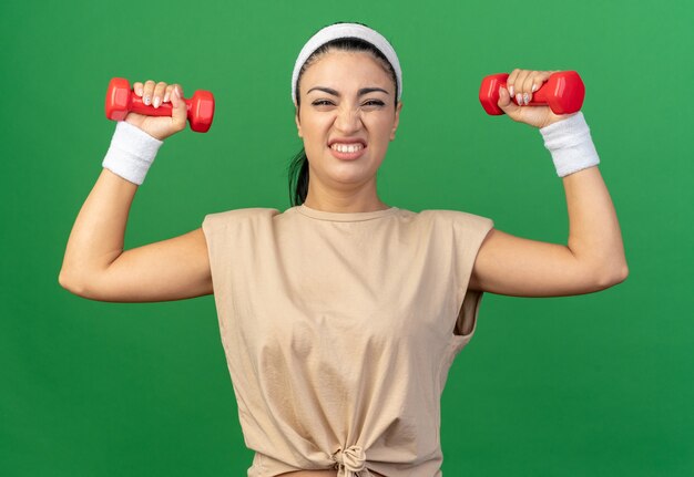 Gespannen jonge kaukasische sportieve vrouw die hoofdband en polsbanden draagt en naar voren kijkt die dumbbells opheft die op groene muur worden geïsoleerd