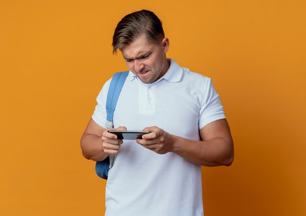 Gespannen jonge knappe mannelijke student die achterzak draagt die op telefoon wordt gespeeld die op oranje wordt geïsoleerd