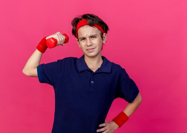 Gespannen jonge knappe sportieve jongen met hoofdband en polsbandjes met beugels hand houden op taille halter verhogen kijken voorzijde geïsoleerd op roze muur met kopie ruimte