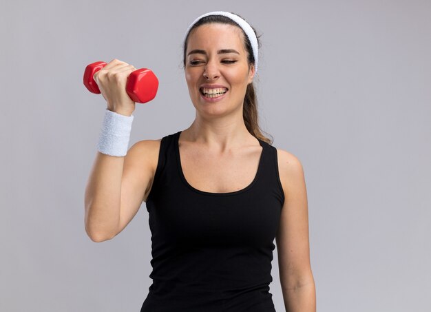 Gespannen jonge, vrij sportieve vrouw met een hoofdband en polsbandjes die een halter opheft en ernaar kijkt