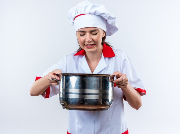 Gespannen jonge vrouwelijke kok die chef-kok uniform draagt die steelpan houdt die op witte achtergrond wordt geïsoleerd