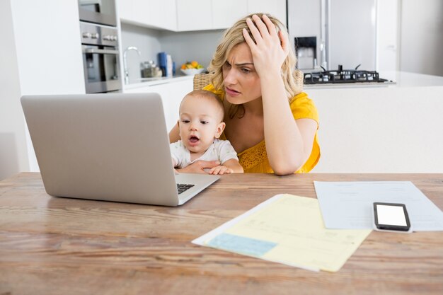 Gespannen moeder met behulp van laptop met haar baby jongen in de keuken