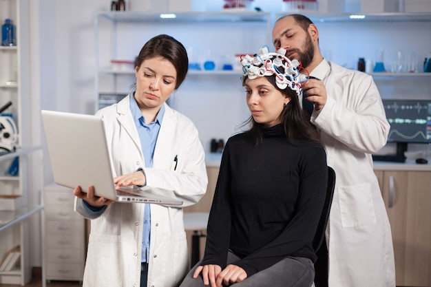 Gespecialiseerde neuroloog arts die aantekeningen maakt op laptop die de symptomen van de patiënt vraagt en de high-tech eeg-headset aanpast. Arts-onderzoeker die EEG-headset bestuurt en hersenfuncties en gezondheidsstatus analyseert.