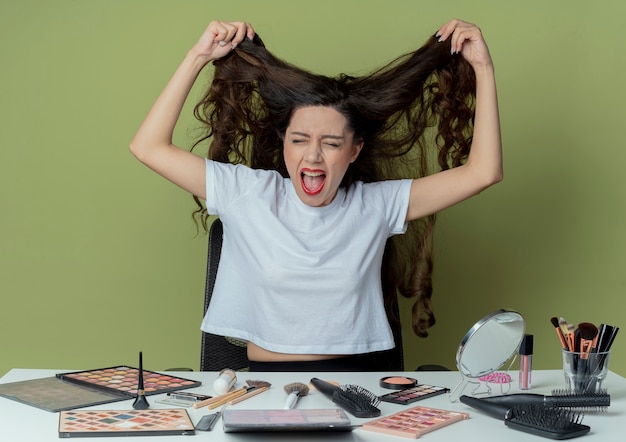 Gestresst jong mooi meisje zittend aan make-uptafel met make-upgereedschap dat haar haar met gesloten ogen op olijfgroene ruimte trekt pulling