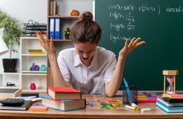 gestresste jonge vrouwelijke wiskundeleraar die aan het bureau zit met schoolbenodigdheden die lege handen laten zien die schreeuwen met strak gesloten ogen in de klas