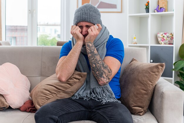 Gratis foto gestresste jonge zieke man met sjaal en wintermuts zittend op de bank in de woonkamer en houdt de handen op het gezicht met servet met gesloten ogen