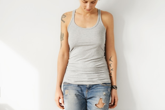 Getatoeëerd jong europees vrouwelijk model neer te kijken op grijs leeg a-shirt. bijgesneden portret van teenge vrouw gekleed in gescheurde jeans en tank top poseren binnenshuis. jeugd en mode