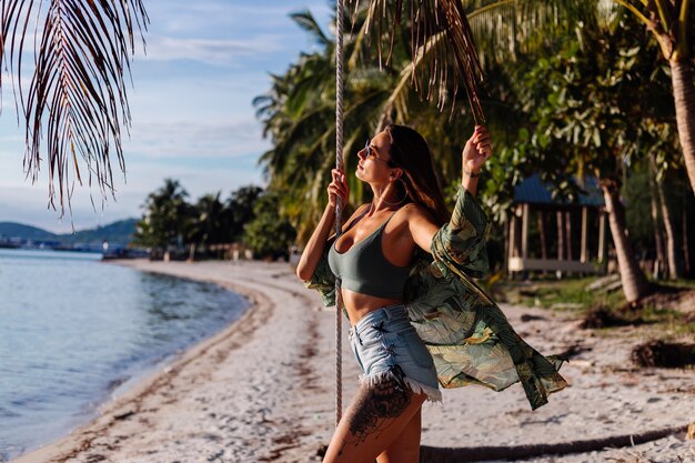 getatoeëerde blanke vrouw in denim shorts en groene trendy crop top op het strand