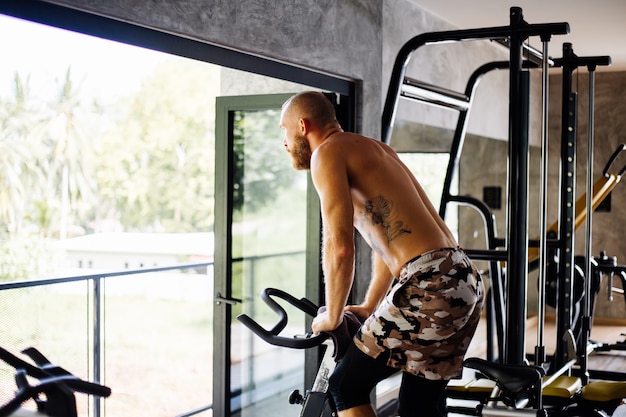 Getatoeëerde gespierde sterke bebaarde man trainen cardio op de fiets in de sportschool in de buurt van groot raam met uitzicht op bomen buiten