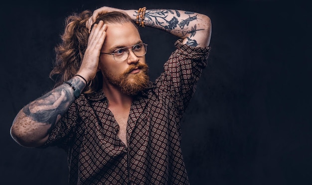 Getatoeëerde roodharige man hipster corrigeert zijn weelderige haar gekleed in een bruin shirt, staande in een studio. Geïsoleerd op de donkere achtergrond.