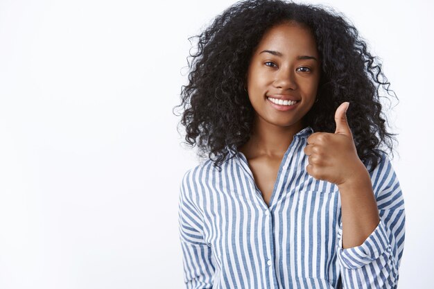 Geweldig goed gedaan. Blij tevreden ondersteunende vrouwelijke donkerhuidige vriend die gestreepte blouse draagt die bevestigend glimlacht en duim omhoog laat zien zoals je idee goedkeuren, grijnzend tevreden witte muur