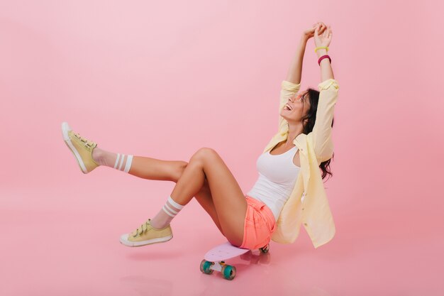 Geweldig Latijns-meisje in roze gestreepte sokken genieten van het leven en glimlachen. Sierlijke jonge vrouw met donker haar poseren met handen omhoog, zittend op een skateboard