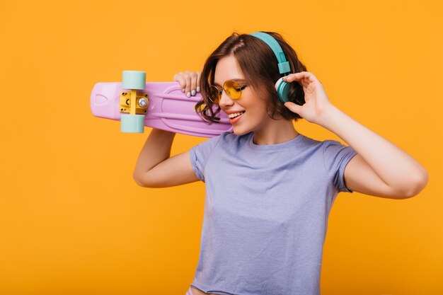 Geweldig meisje in blauwe koptelefoon dansen. Binnen schot van enthousiaste jonge vrouw die met skateboard geluk uitdrukt.
