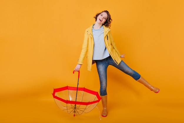 Geweldig meisje in blauwe spijkerbroek en shirt grappig dansen, paraplu te houden en glimlachen. vrolijke dame in herfst jas en rubberen schoenen plezier na regen.