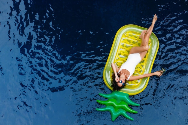 Geweldig meisje in zonnebril liggend op ananas matras. Buiten foto van mooi gelooid vrouwelijk model in zwembroek ontspannen in het zwembad.