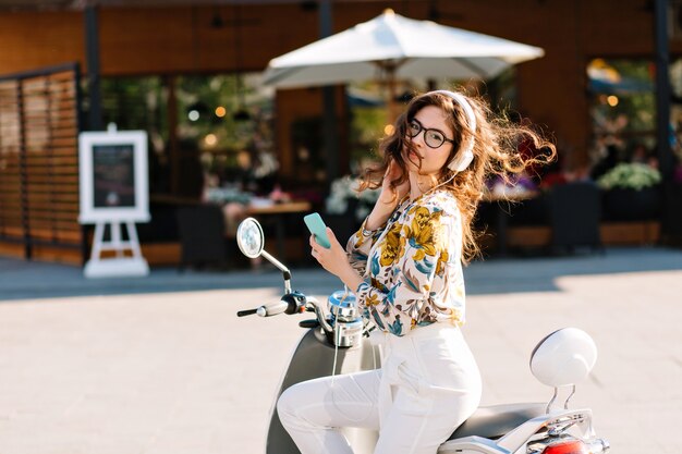 Geweldig stijlvol meisje met telefoon op scooter te wachten op vriendje die voor koffie ging in café