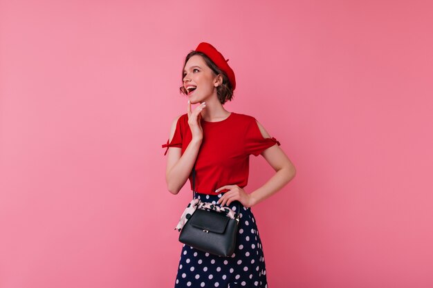 Geweldig vrouwelijk model in Franse outfit poseren met opgewonden gezichtsuitdrukking. Indoor portret van geïnteresseerde Europese vrouw met zwart lederen handtas.